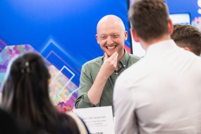 Man with green shirt smiling as he speaks to 3 people in front of a desk at an exhibit at the Esri UK Annual Conference 2026.