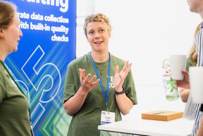 three people standing in front of banner and desk at the Esri UK Annual Conference 2025 with woman in the middle explaining something