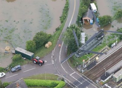 Aerial view of a flooded rural area with a military helicopter flying overhead, surrounded by submerged fields, a railway line, and vehicles navigating partially flooded roads.