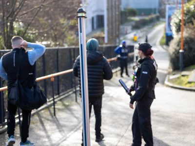 3 police in uniform and undercover facing away standing near metal detector