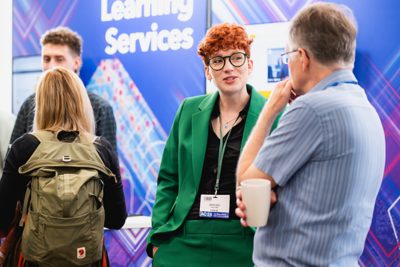 Three people at the Esri UK Annual Conference 2025 talking in front of an exhibit, with the main focus on a man with a green shirt and glasses in the middle