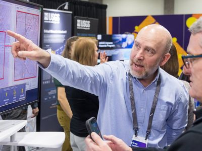 Attendee in a blue dress shirt wearing a lanyard points to a screen showing maps