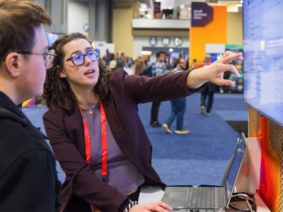 Esri expert wearing a red lanyard pointing at a screen and talking to an attendee at an expo booth