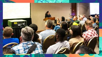 Audience seated in rows facing a speaker at a podium presenting slides in a conference room