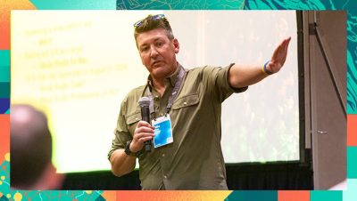 Speaker holding a microphone and gesturing toward a presentation slide behind them