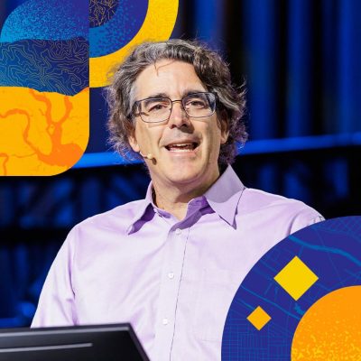 Presenter in a purple shirt and glasses speaking onstage, bordered by artwork of geometric shapes containing map elements