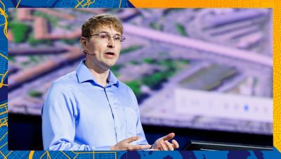 Presenter speaking onstage with a photograph of city structures in the background