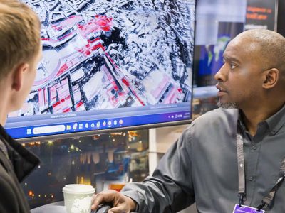 Exhibitor and attendee look at a map displayed on a large monitor in an exhibitor’s booth