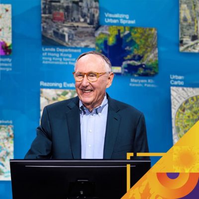Esri president Jack Dangermond onstage with a display of maps in the background
