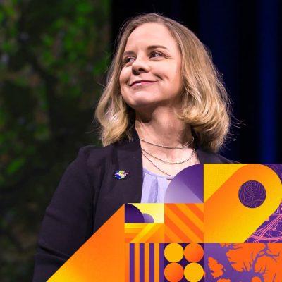 Blonde presenter in a black jacket looks to the left and smiles; shapes in yellow, purple, and orange are underneath