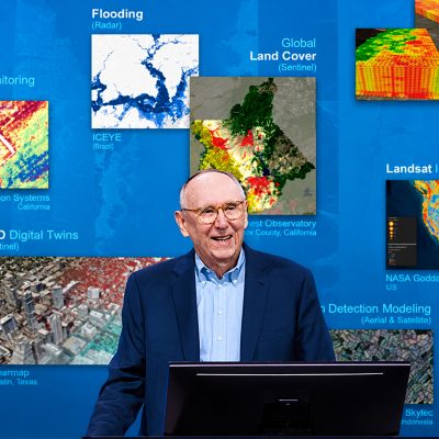 Esri president Jack Dangermond onstage with a display of maps in the background