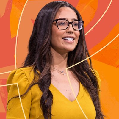 A woman with long dark hair wearing a yellow sweater and glasses smiling as she speaks