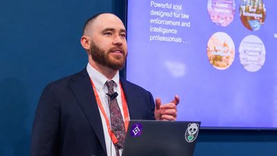 Presenter wearing a suit and tie looks out at the audience with a screen behind him