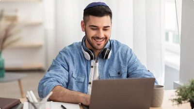  A man smiles as he works on a laptop at a desk while wearing headphones around his neck