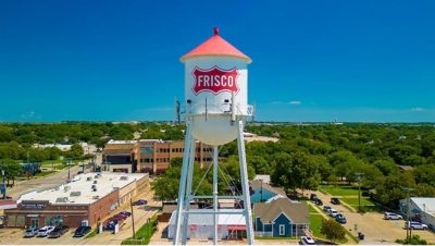 A white water tower with “Frisco” printed on it standing tall above a peaceful tree-filled town under a blue sky