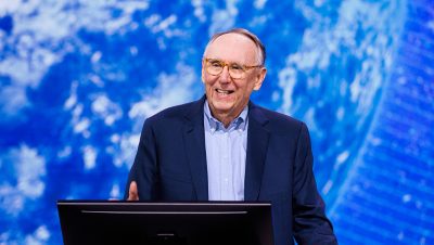Jack Dangermond wearing a blue suit mid-speech at a podium with a blue graphic in the background