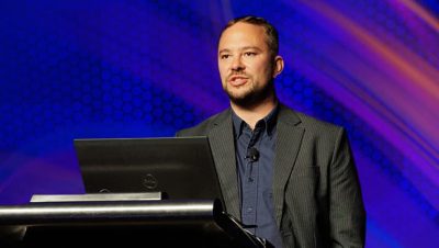Presenter speaking at a podium against a blue background with subtle red swirls