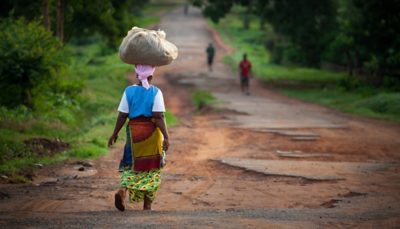 Person walking down a partially paved road in a forest carrying a tied-up sack on their head