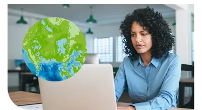 A woman wearing a blue shirt is seated at a desk in a modern office environment, working on a laptop. 