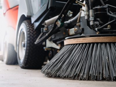 Low angle close-up of a street sweeper showing the large circular mechanical brush and tires