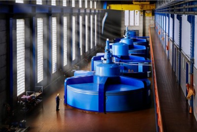 A large blue turbine sits in a room with large windows. A worker in a yellow hard hat stands next to it.