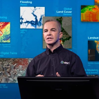 Presenter wearing a black button-up shirt with an embroidered Esri logo in front of a screen displaying user image submissions
