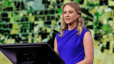 Presenter in a blue top speaking onstage with a black and green map in the background