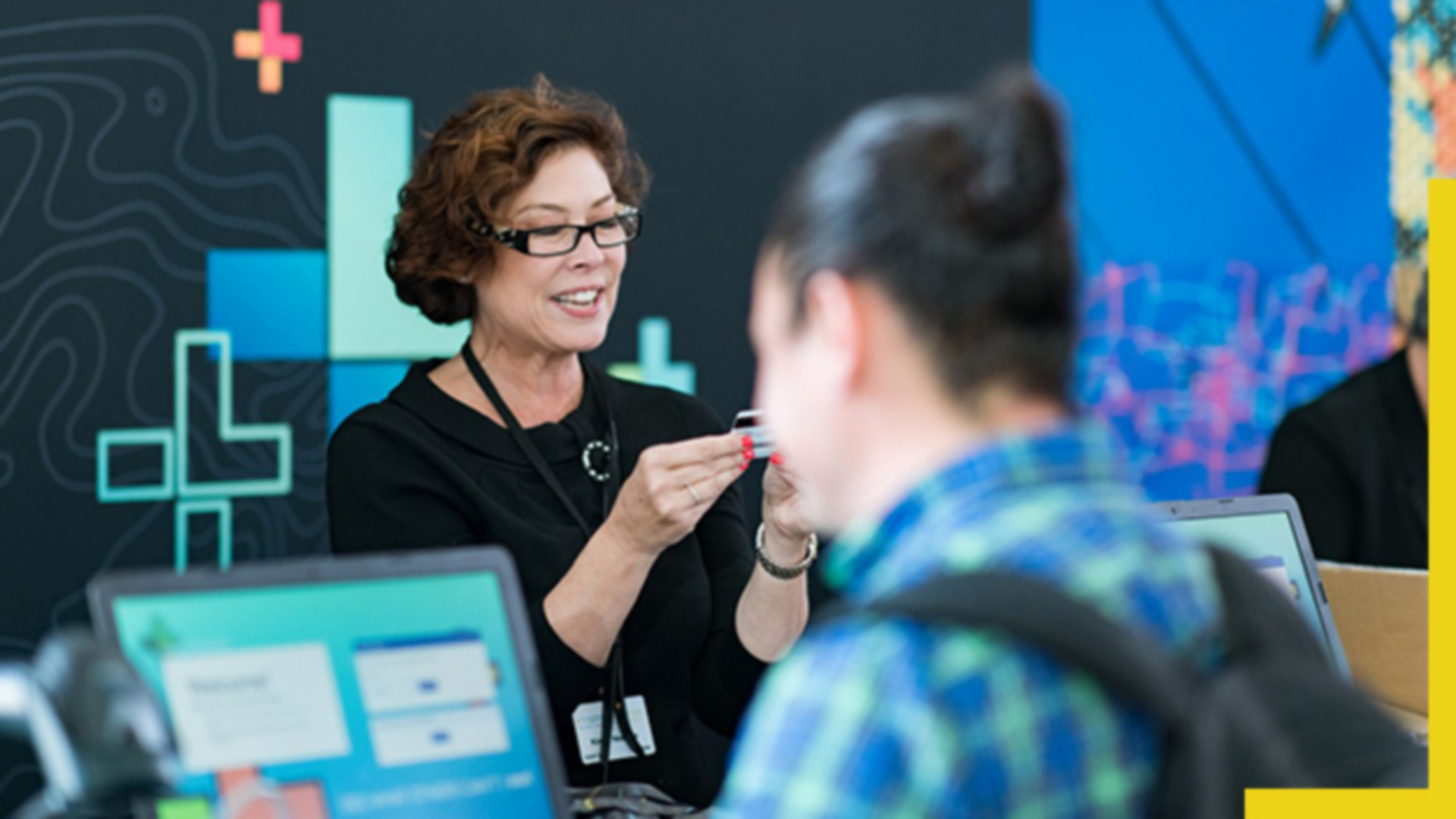 A photo of a conference staffer helping a registrant while a person wearing a backpack works on a laptop in the foreground 