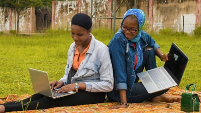 Two people sit back-to-back on a patterned blanket in a grassy outdoor area, each using a laptop computer. 