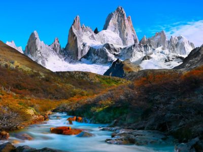 A dramatic range of jagged, snow-covered mountains rises sharply under a clear blue sky. 