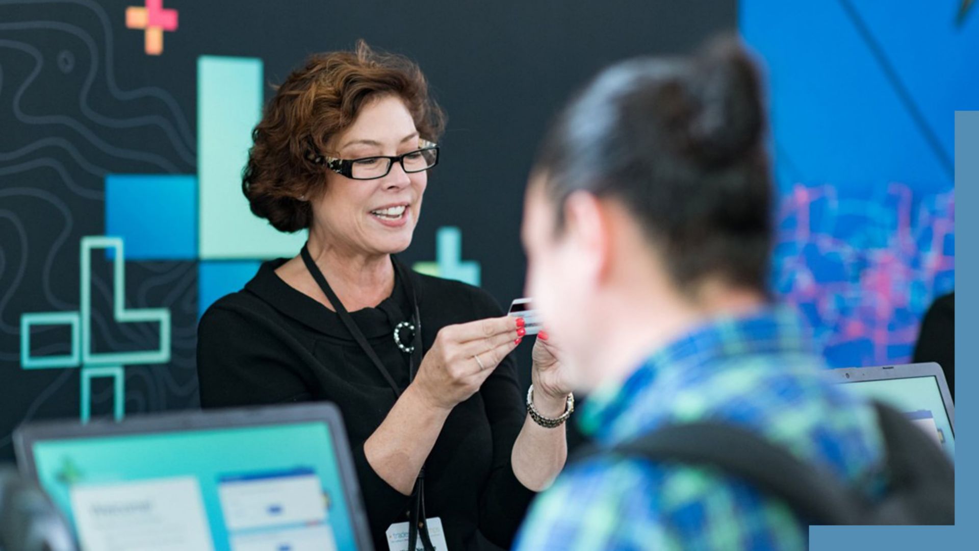 A photo of a conference staffer helping a registrant, while a person in a backpack stands in the foreground using a laptop