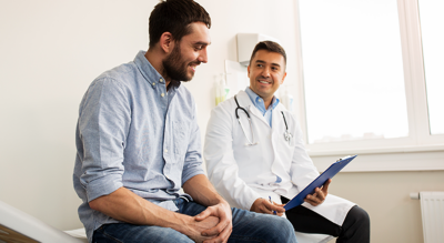 A male patient sits attentively while a doctor in a white coat holds a clipboard during a consultation.