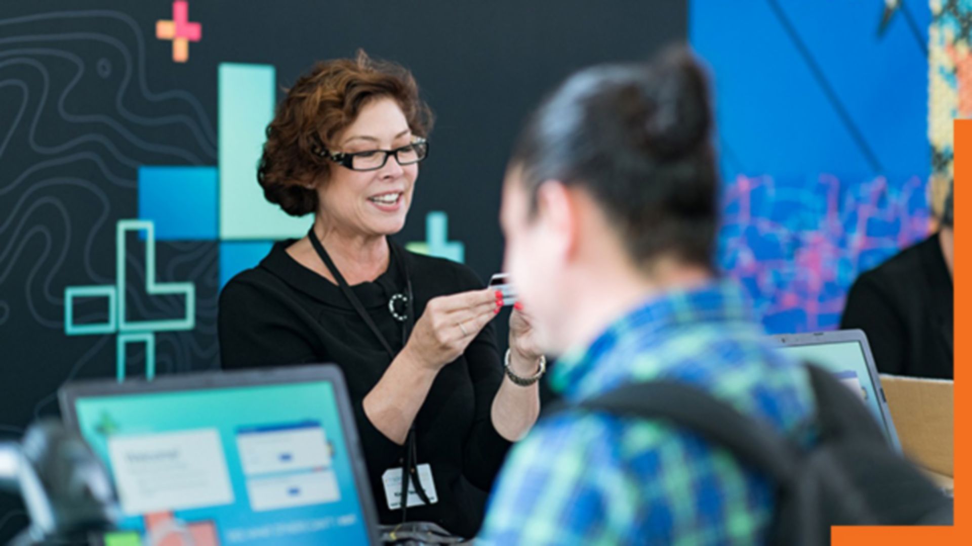 A photo of a conference staffer helping a registrant, while a person in a backpack stands in the foreground using a laptop