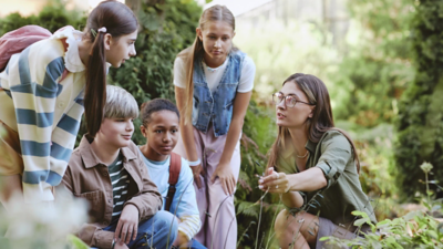 A group of students standing in a sunlit forest listening intently to a teacher as she speaks and gestures at nearby foliage