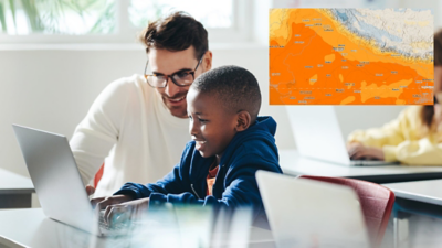 Ein Lehrer hilft einem lächelnden Schüler an einem aufgeklappten Laptop in einem hellen, modernen Klassenzimmer, überlagert mit einer bunten Weltkarte