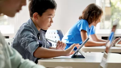 A row of students sitting at their desks using tablets in a sunlit modern classroom