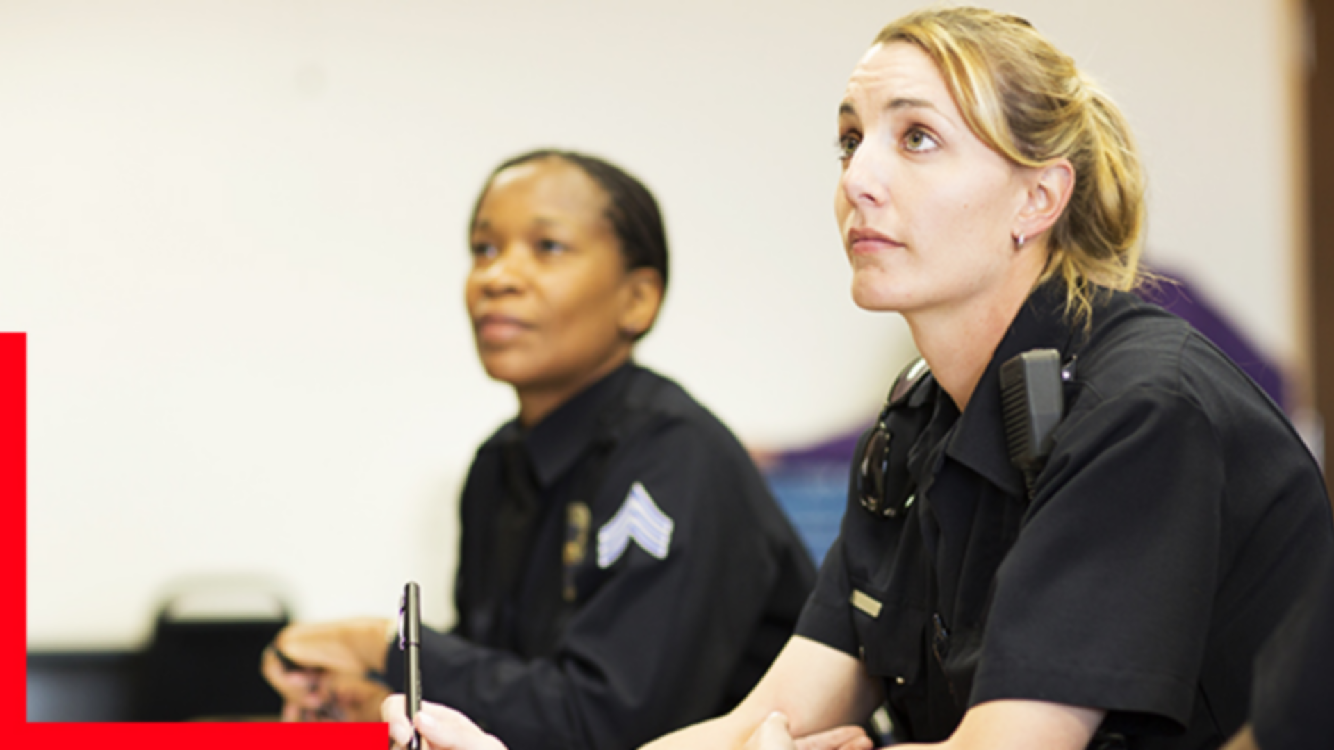 Two police officers in uniform sit at desks, listening attentively.