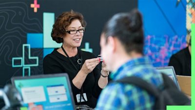 A photo of a conference staffer helping a registrant, while a person in a backpack stands in the foreground using a laptop