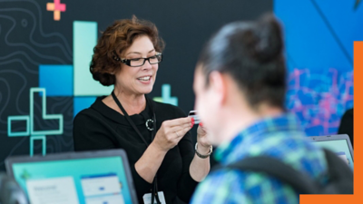 A photo of a conference staffer helping a registrant, while a person in a backpack stands in the foreground using a laptop