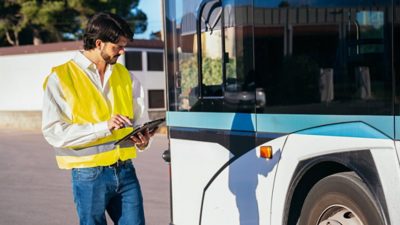A worker wearing a safety vest using a tablet to inspect the outside of a bus