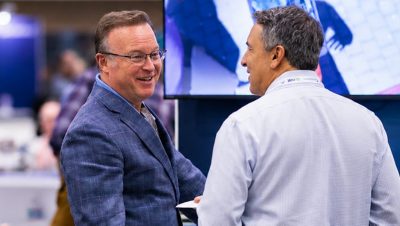 Two middle-aged men in business attire converse in a professional conference setting.