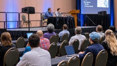 A group of attendees sit at tables listening to two speakers at a conference panel discussion.