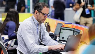 Person working on a laptop at an event seated at a shared table with other attendees and colorful booths in the background