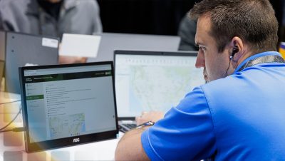  Person wearing a blue polo shirt and ear buds working on a laptop that shows step-by-step directions and a map