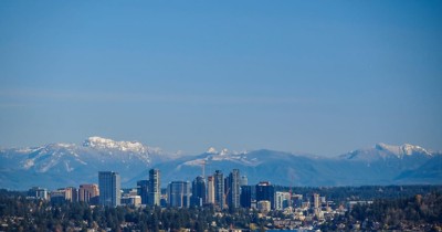 Urban skyline with high-rise buildings set against snow-capped mountains and a clear blue sky.