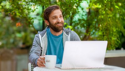 A man sits at an outdoor table with a laptop and a white coffee mug featuring the text 'Esri'. 