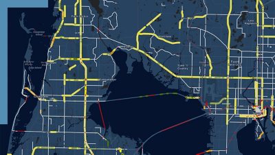A dark-themed map showing a roadway network with color-coded traffic conditions and route lines around a lake