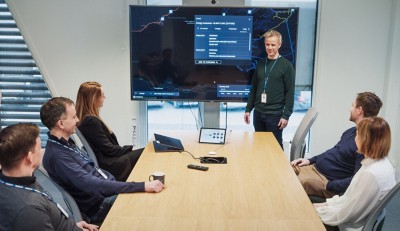 Modern office meeting: six individuals seated at a table, facing presenter and a large screen displaying a map and data.