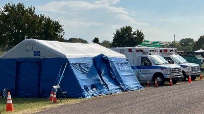 A large blue medical tent with two ambulances parked beside it and trees behind it