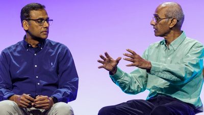 Two seated speakers on stage in front of a purple gradient backdrop